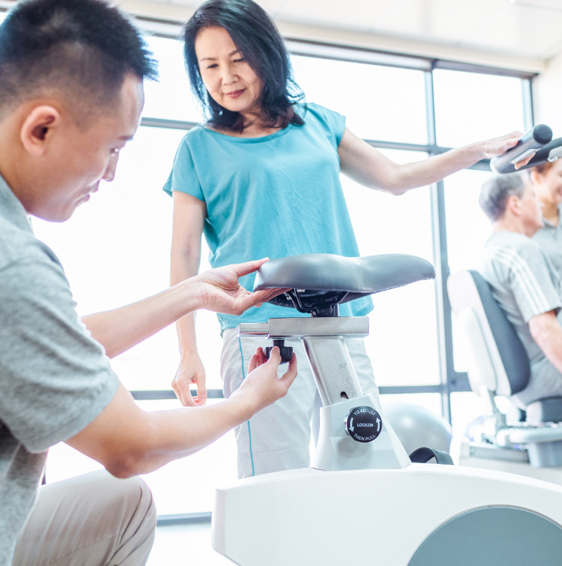 Dyaco Medical Physical Therapy Upright Bike 4.0U having its seat height adjusted by therapist while patient looks on.