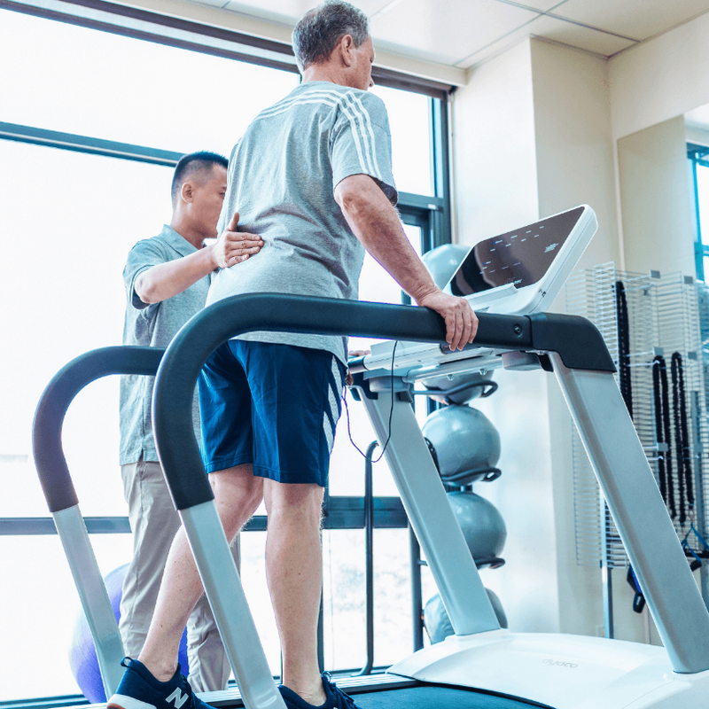 Dyaco Medical Physical Therapy Treadmill 4.0T in gym room with male patient viewed from behind with the therapists hand placed in the patients lumbar region of his back.