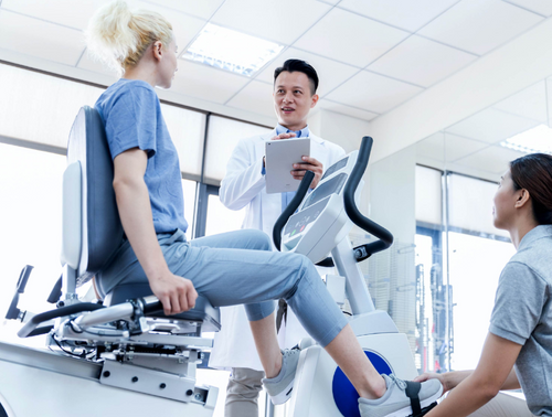 Dyaco Medical Rehabilitation 7.0R Recumbent Bike in gym room with female patient on the bike with one therapist helping put her shoe into pedal and another taking notes.