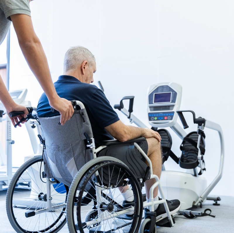 Dyaco Medical Rehabilitation Recumbent Seated Stepper 7.5S showing therapist pushing patient in wheelchair into wheelchair into position.