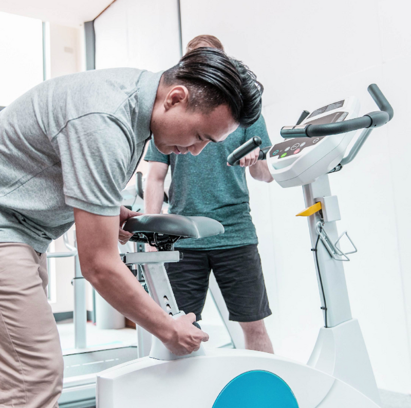 Dyaco Medical Rehabilitation Upright Bike 7.0U in gym room with thearpist altering the seat height with aptient standing at the side of the bike.