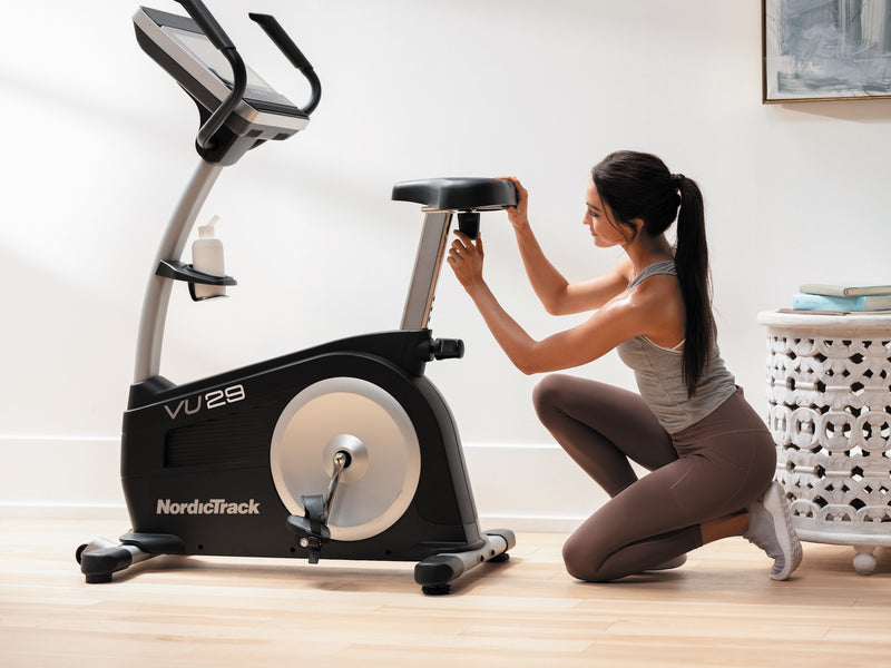Nordic Track VU29 Upright Bike. An image of a female demonstrating how the saddle on the bike moves forwards and backwards.