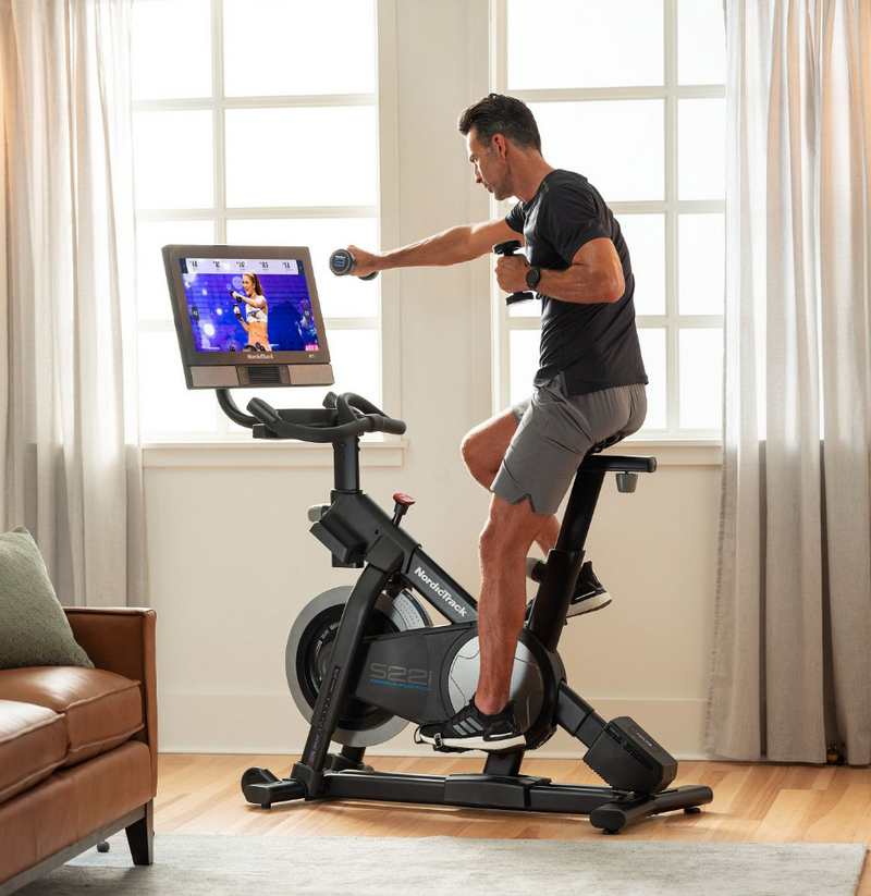 Nordic Track S22i Studio Bike male model using dumbbells sat on bike in room