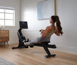 A person using the NordicTrack RW 300 Rower machine, which is a modern rowing machine with a digital display and black color palette, in a home exercise environment.