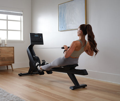 A person using the NordicTrack RW 300 Rower machine, which is a modern rowing machine with a digital display and black color palette, in a home exercise environment.
