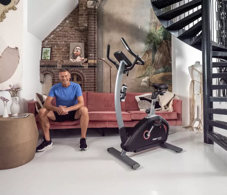 Flow Fitness DHT 2000i Upright Bike with male sitting on sofa in room 